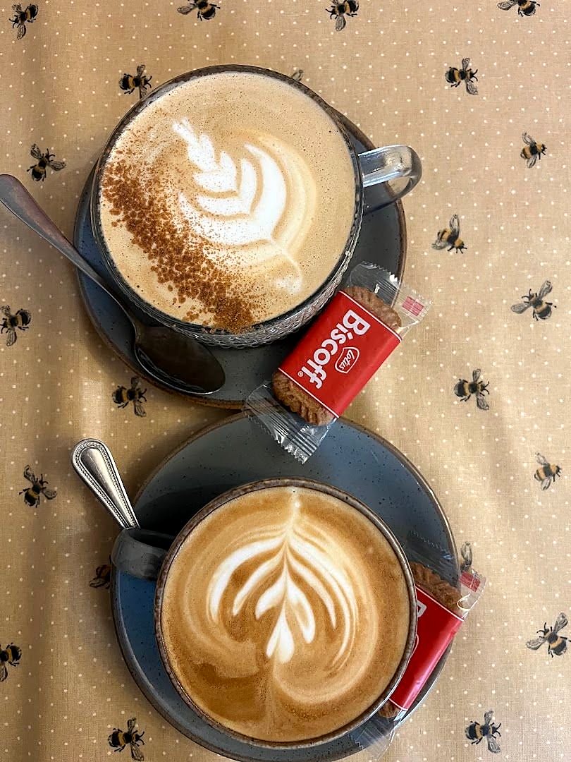 Two lattes with leaf and swan patterns in the foam, each with a wrapped Biscoff biscuit, on a tablecloth printed with little bees
