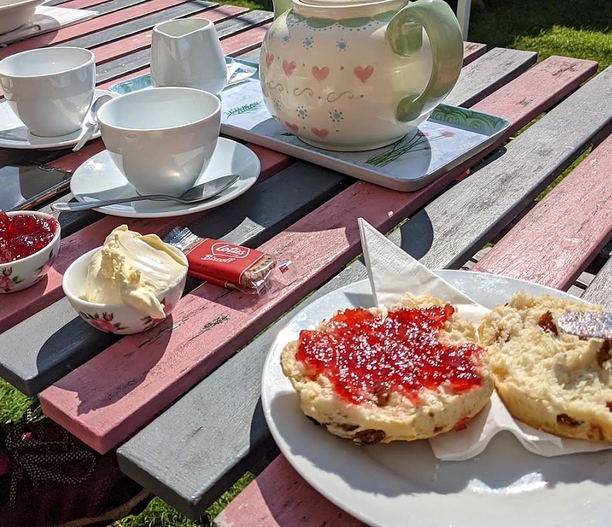 Cream tea at a pink and grey picnic table in the garden: a scone spread with strawberry jam, a pot of clotted cream, a dish of jam, cups and a teapot with pink hearts