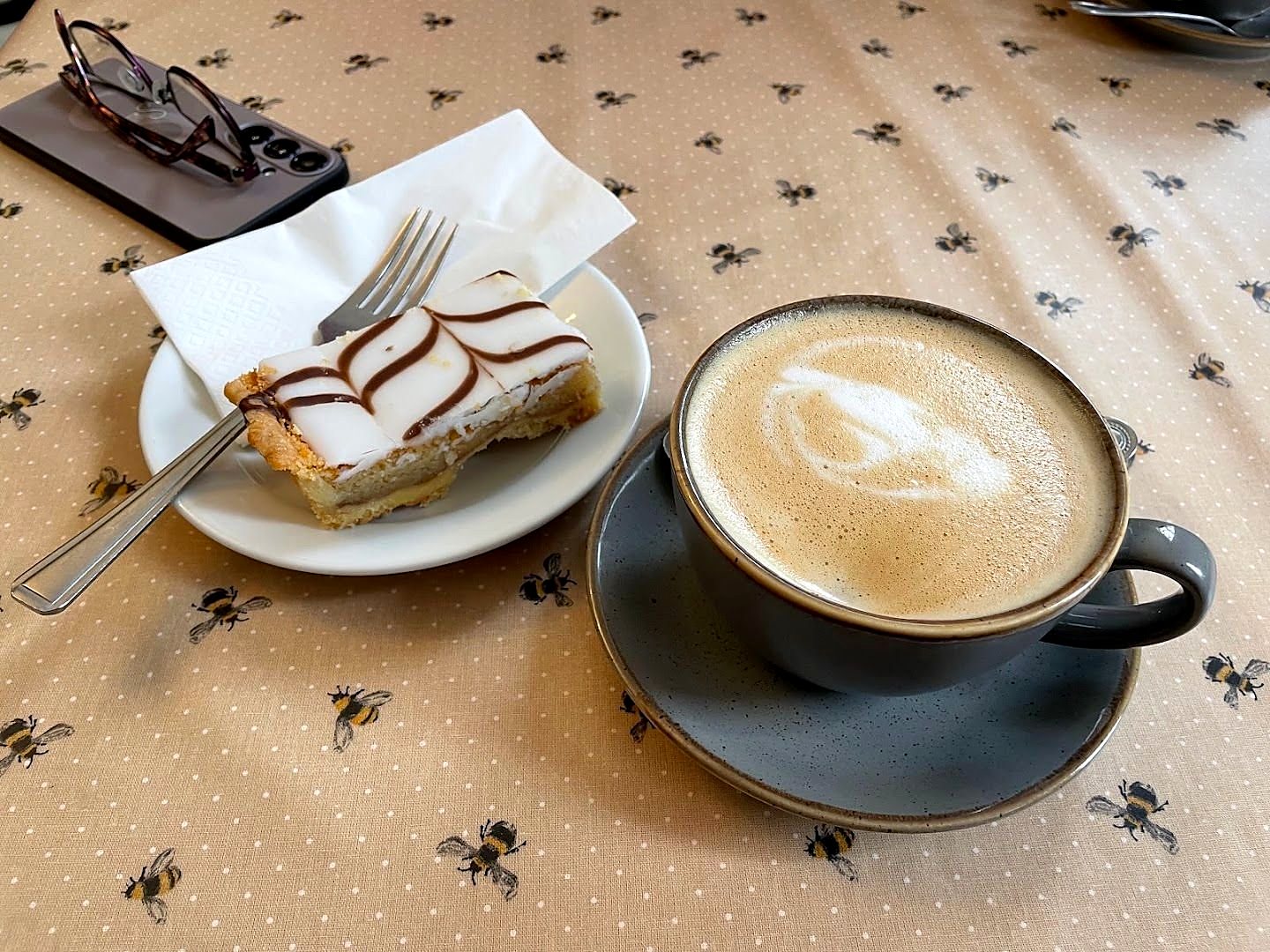 A vanilla slice with feathered icing on a white plate next to a latte, on a tablecloth printed with little bees